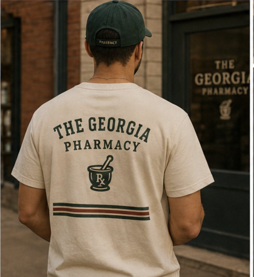 A person in a cream Georgia Pharmacy tee and green cap standing outside the pharmacy storefront